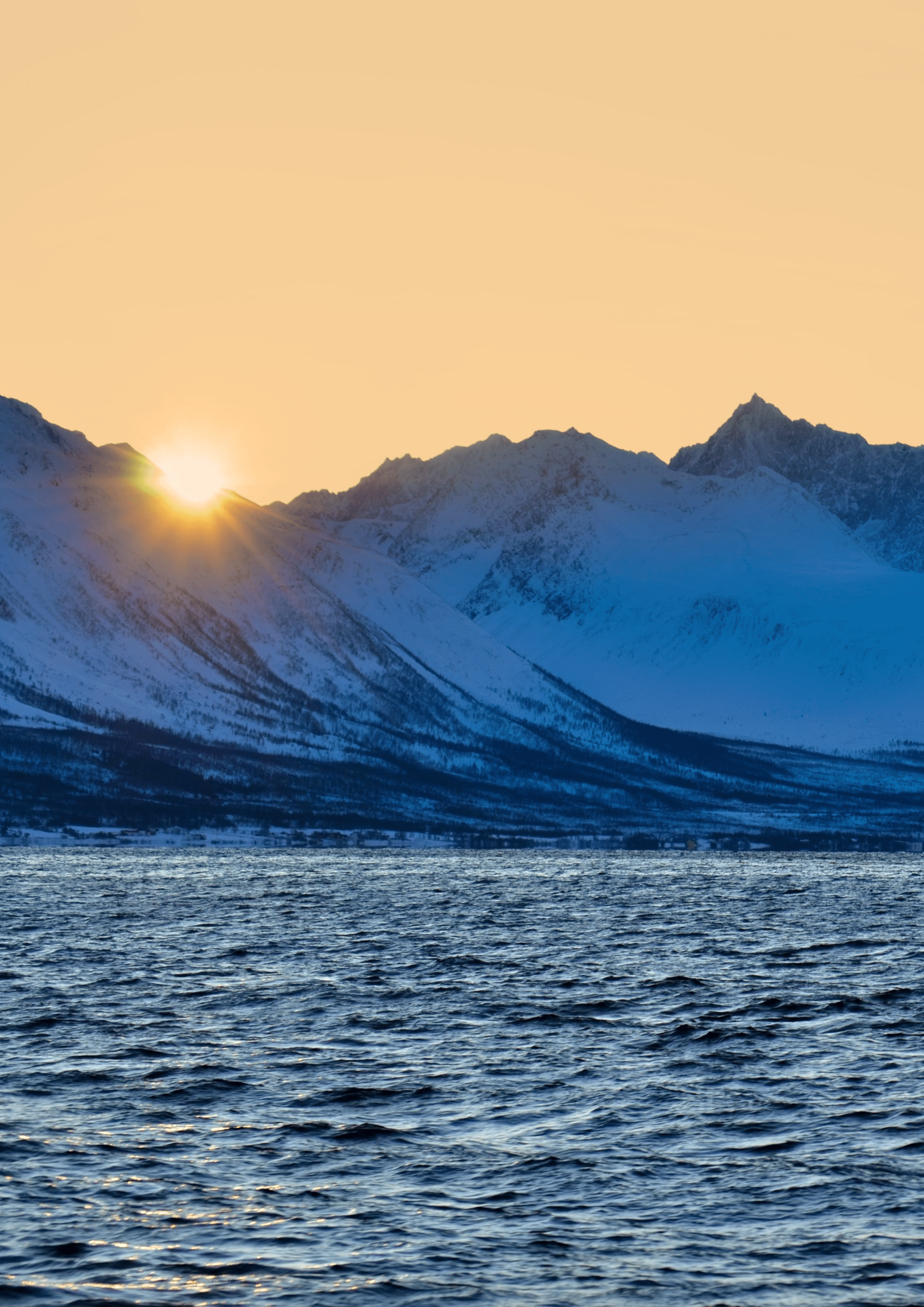 Snowy mountains at sunset over a body of water, sun peaking over the peaks.