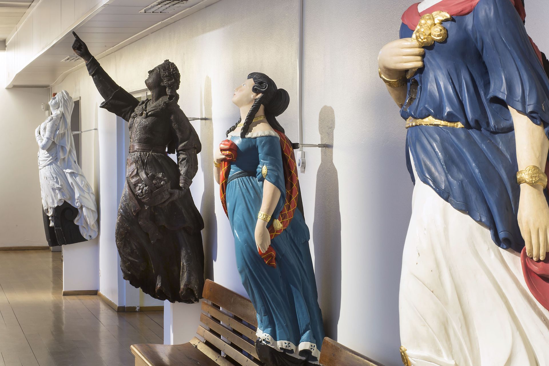 Ship figureheads of women in different colors displayed on a white wall in an art gallery.