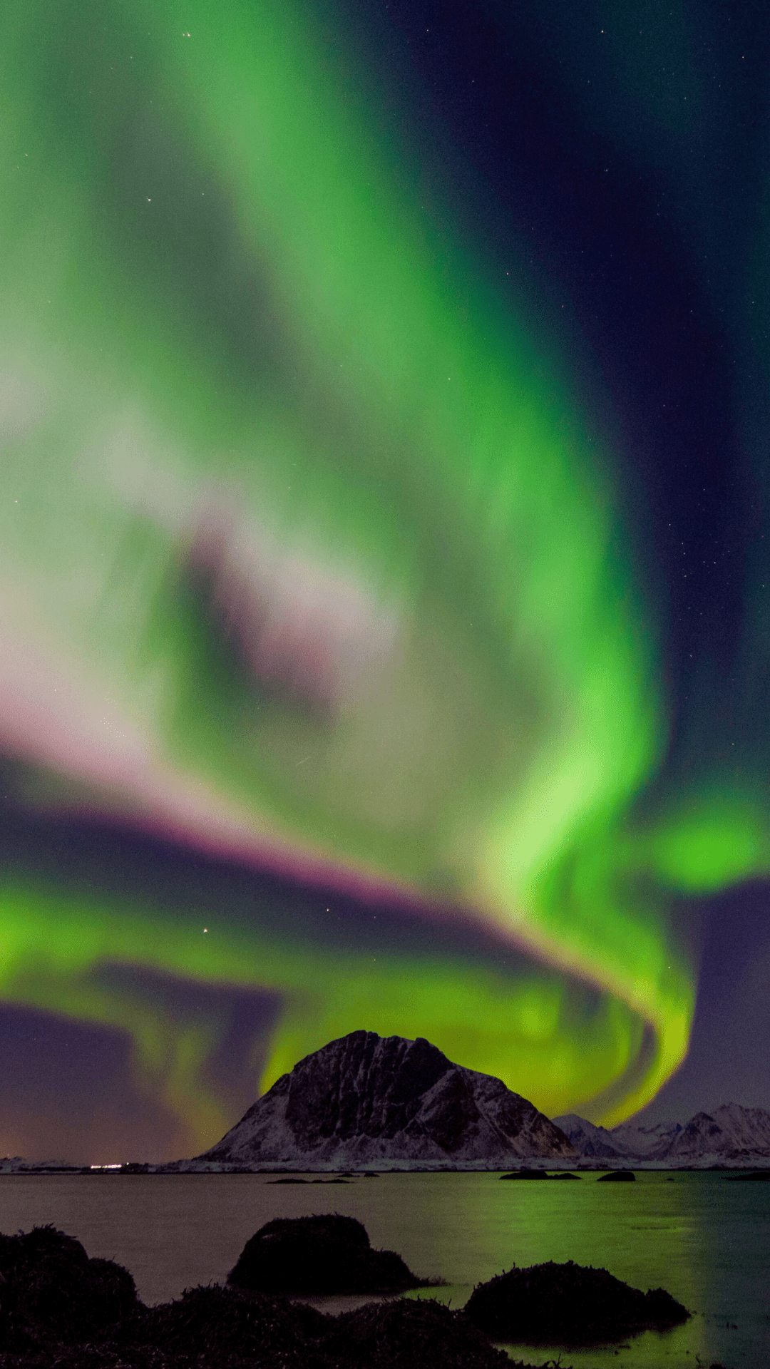Green and purple aurora borealis swirling above dark mountain and water.