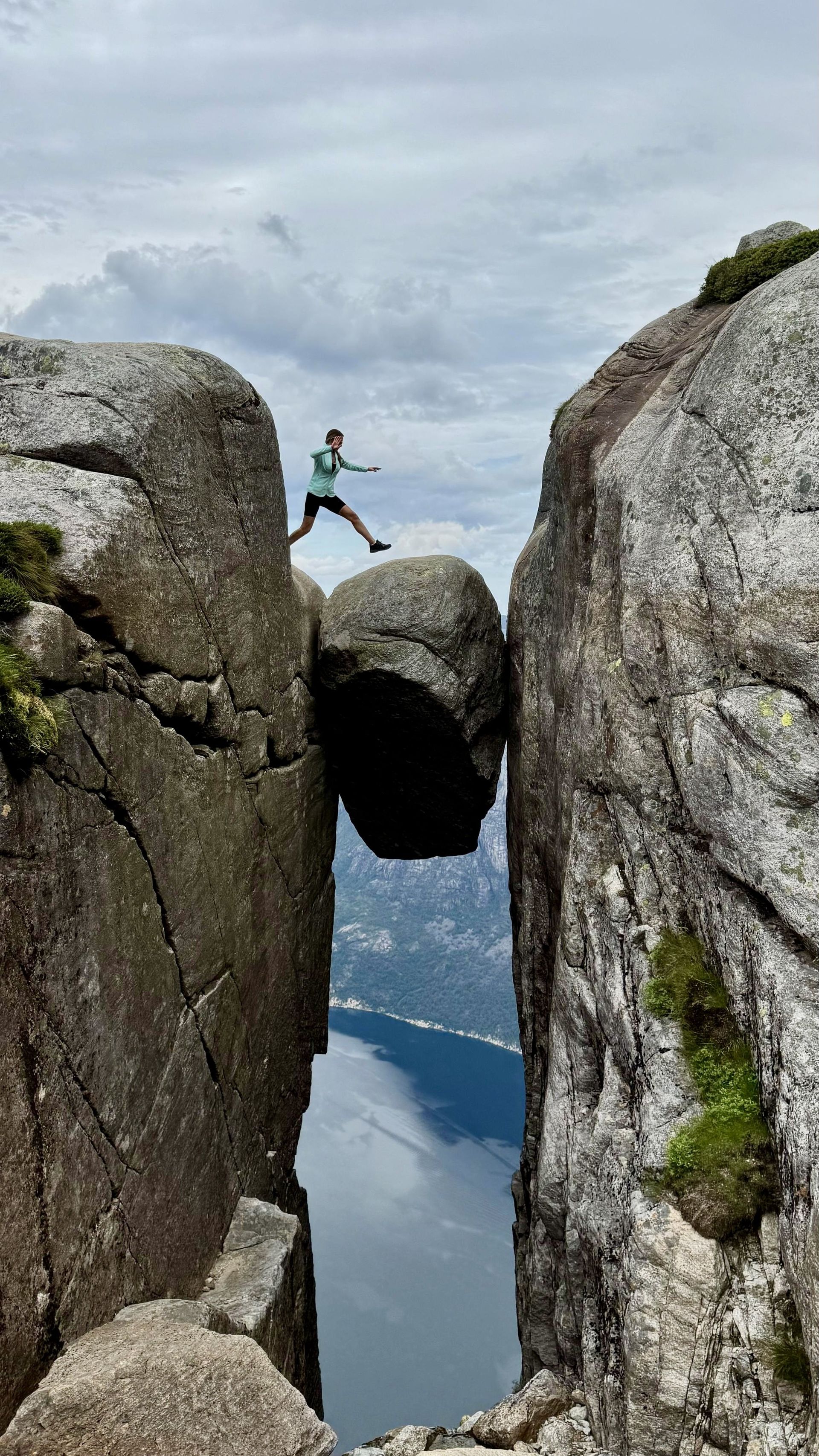 Person jumping across a chasm between large rocks. A balanced boulder sits in the gap. Overcast day, water visible below.