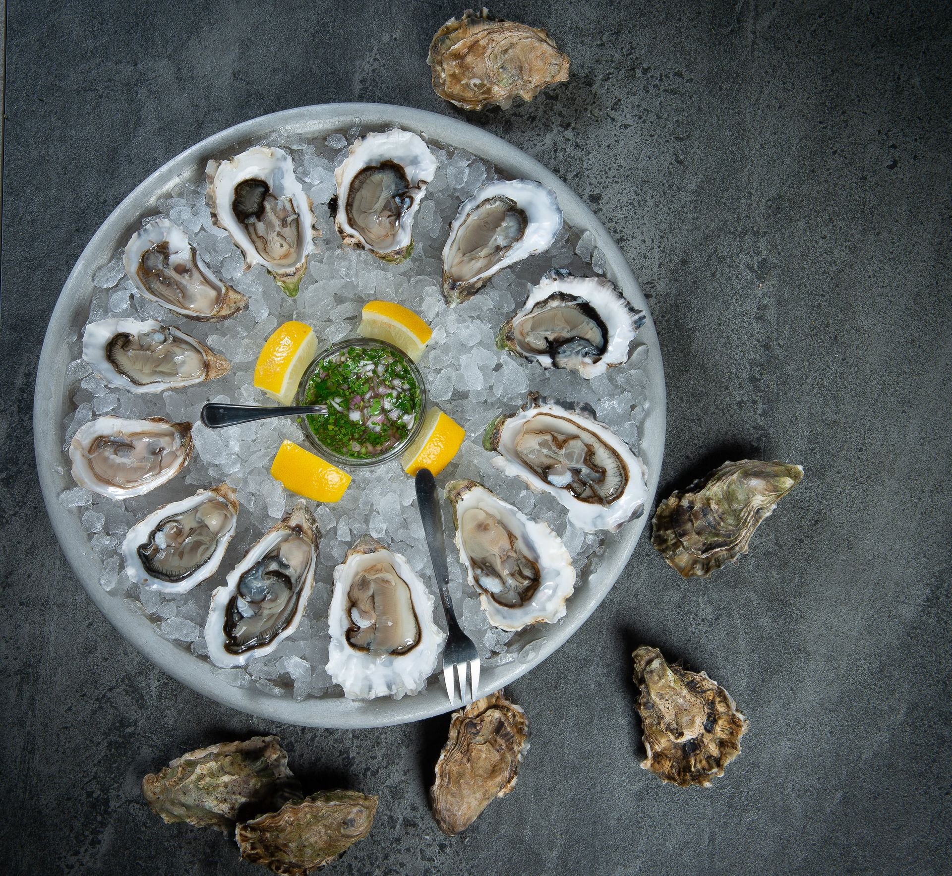 Oysters on ice with lemon wedges and sauce on a gray platter. Additional oysters scattered around.