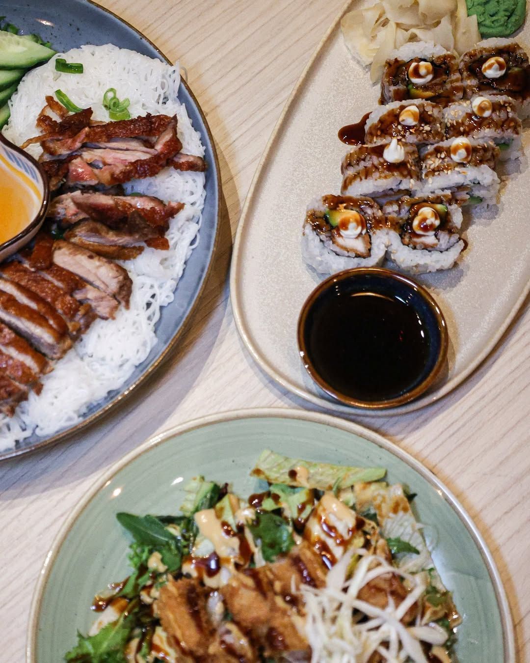 Duck dish, sushi rolls, and salad on plates, served on a light wood table.