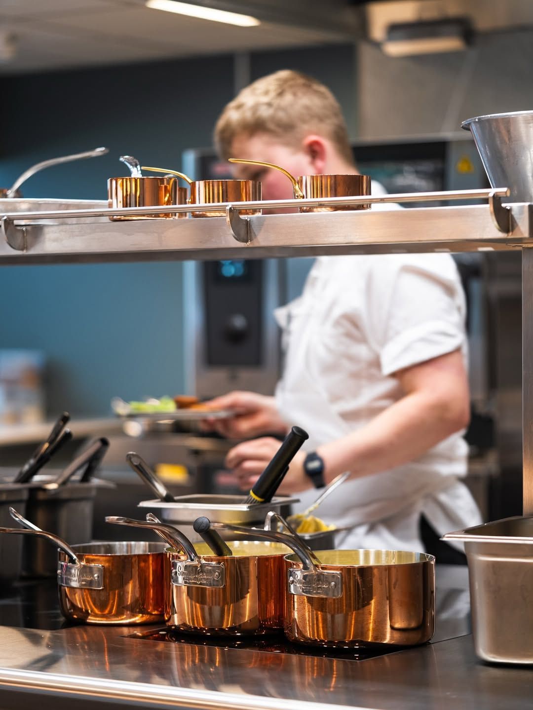 Chef in white uniform cooking with copper pots on a stovetop in a kitchen.