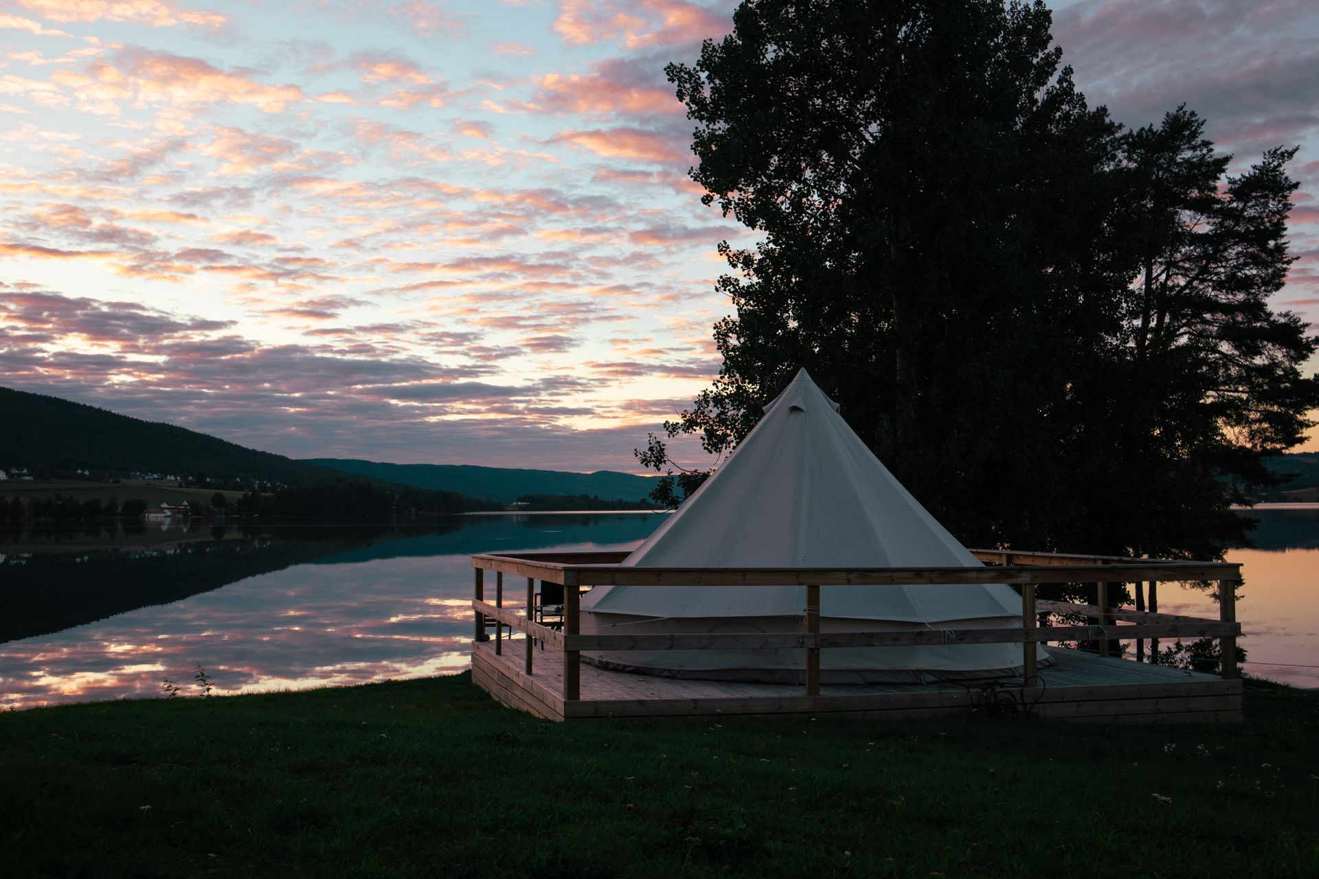 White tent on wooden deck by a lake at dusk, with mountains and colorful sky.