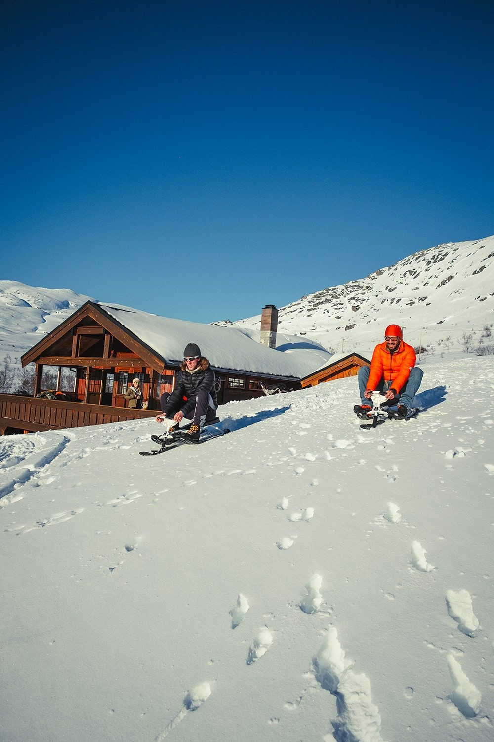 Two people on sleds slide down a snowy hill towards a wooden cabin on a sunny day.