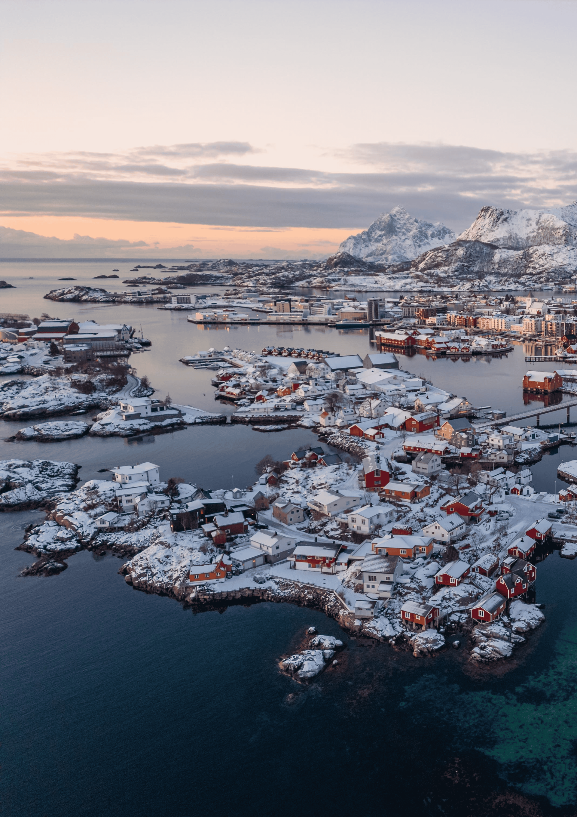 Snow-covered village on islands in Norway, with mountains in the background. Red and white buildings, blue water.