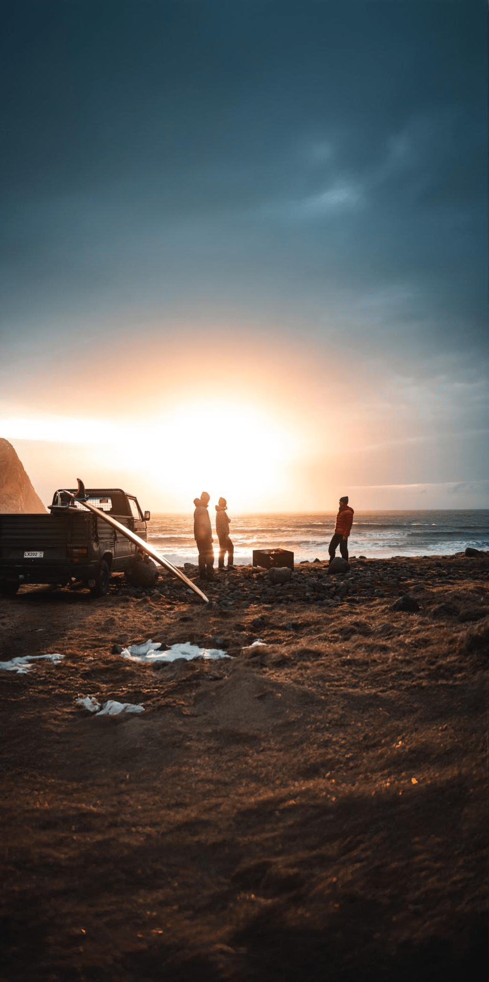 Truck and people silhouetted on a beach at sunset, with ocean and dark clouds.