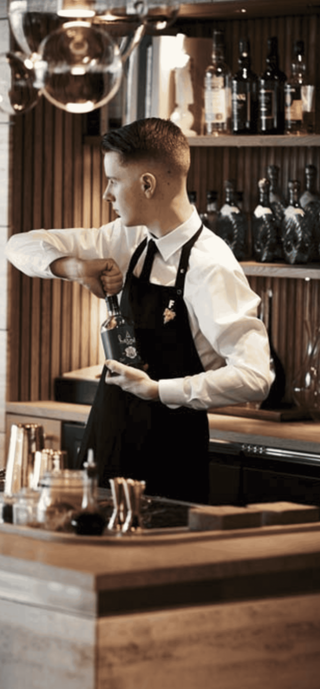 Bartender in a white shirt and black apron, opening a bottle at a bar.