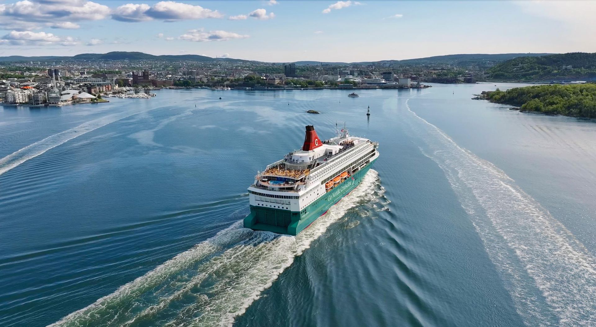 Cruise ship sailing through a blue harbor, leaving a white wake near a coastal city.