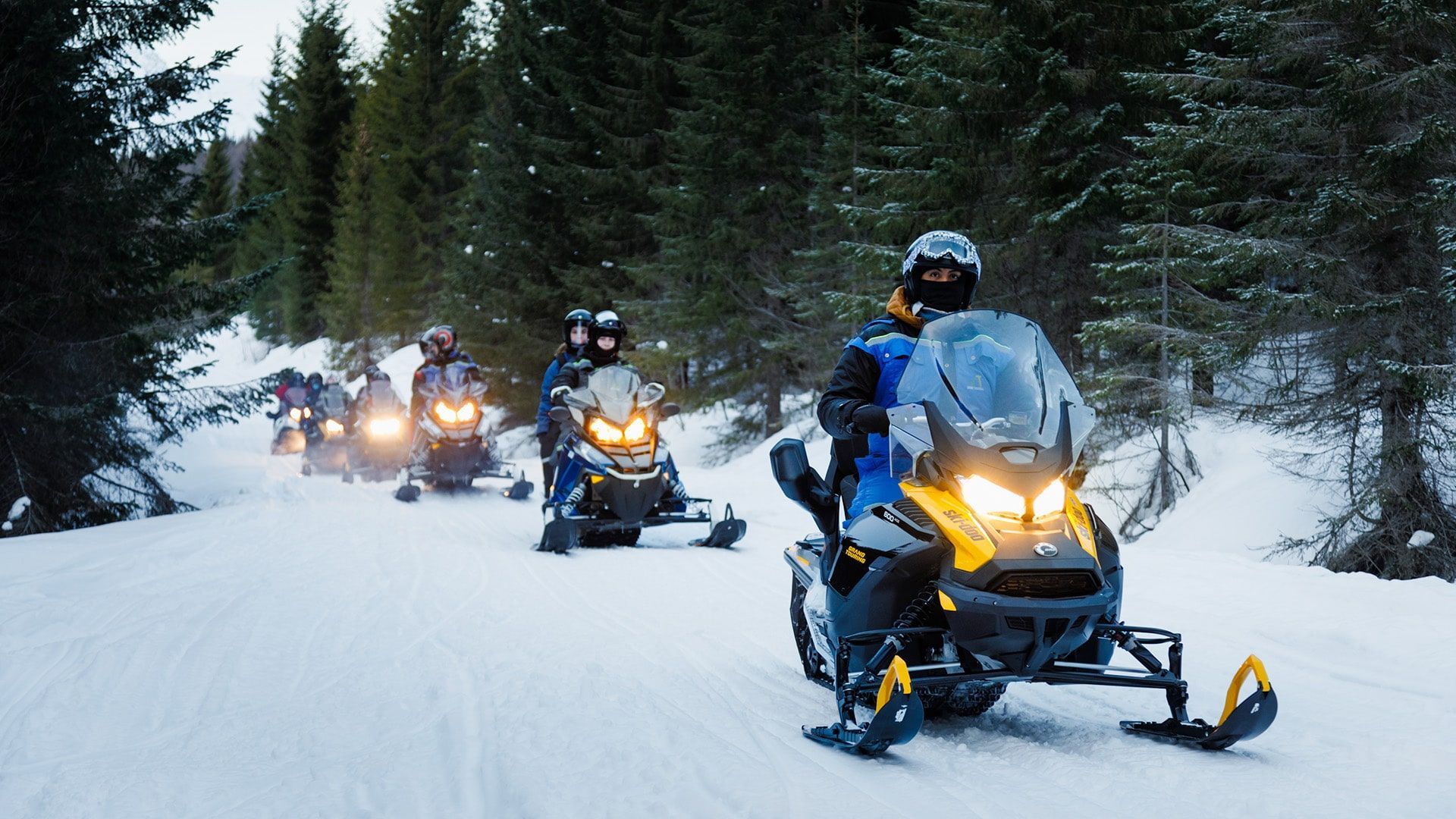 Group of people snowmobiling on a snowy trail through a forest.