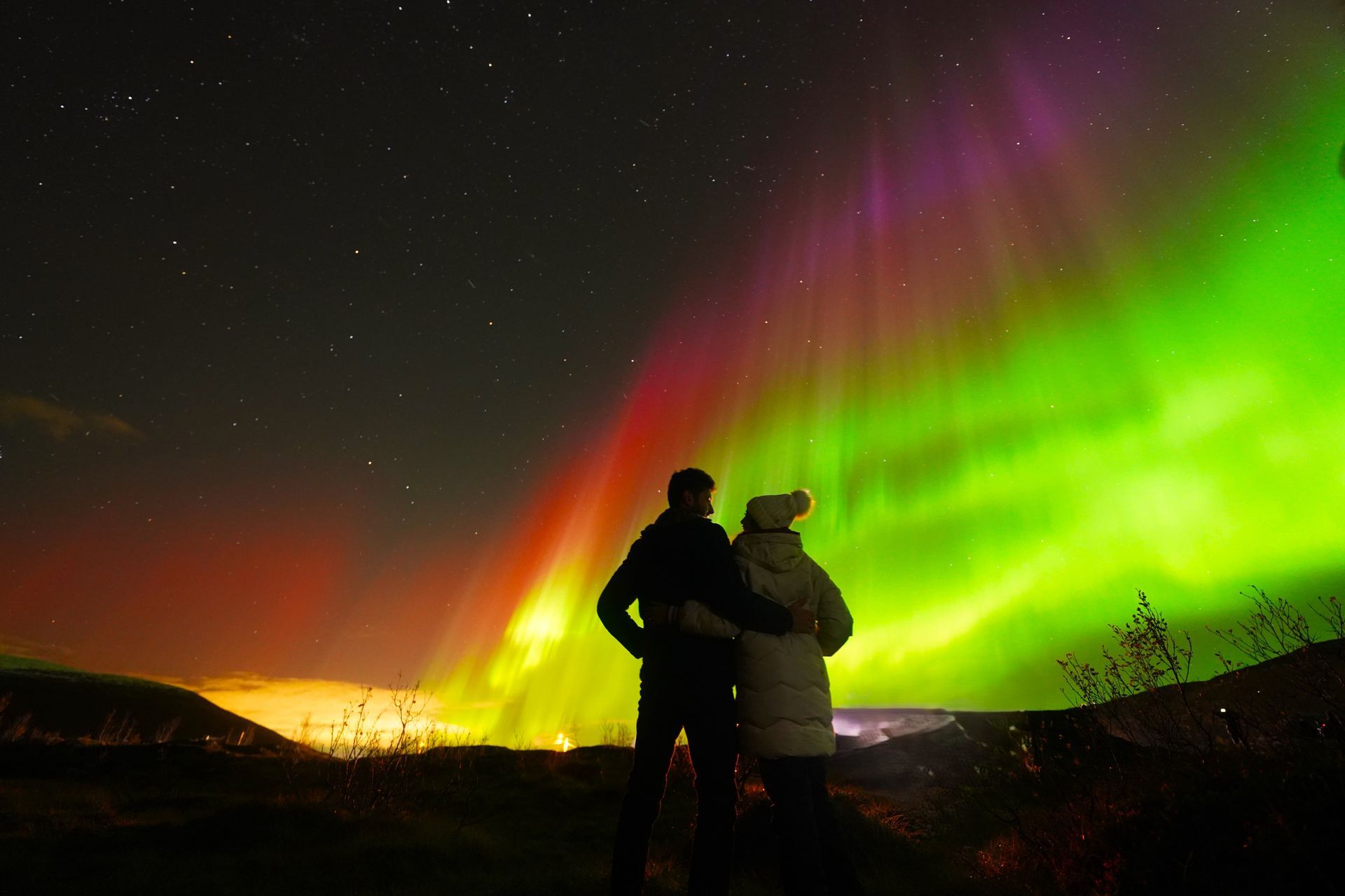 Couple watches vibrant green, purple, and red aurora borealis in a dark night sky.