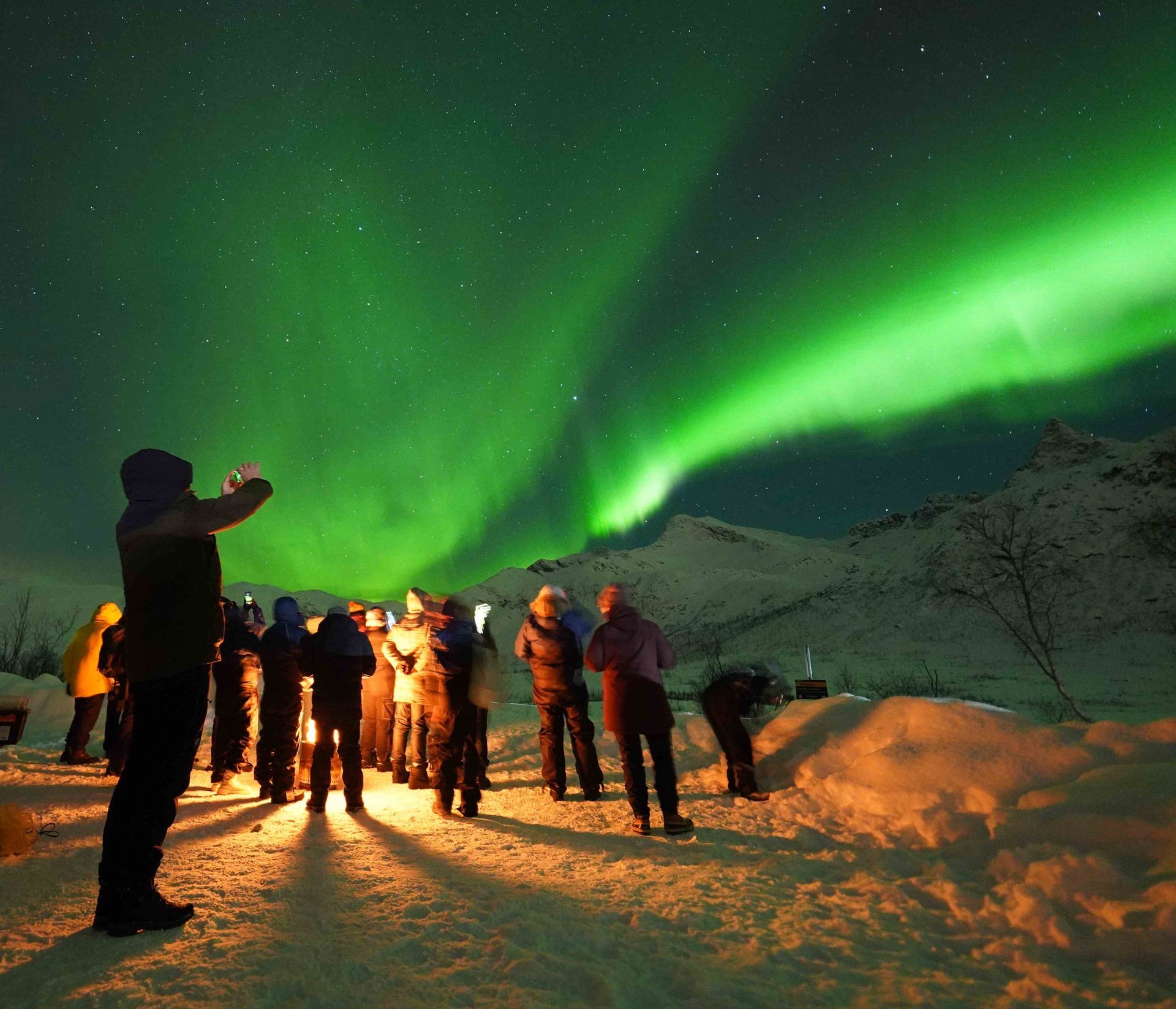 People watch the Northern Lights, green aurora borealis, over snow-covered mountains at night.