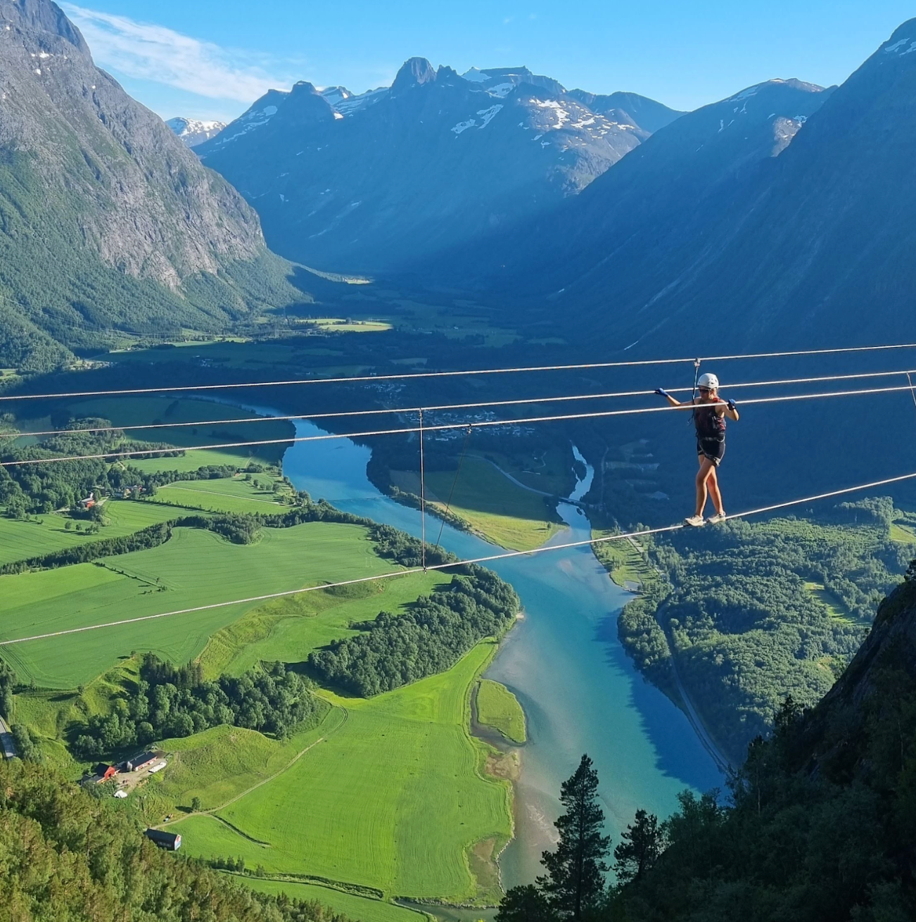 Person tightrope walking over a scenic river valley, surrounded by mountains.