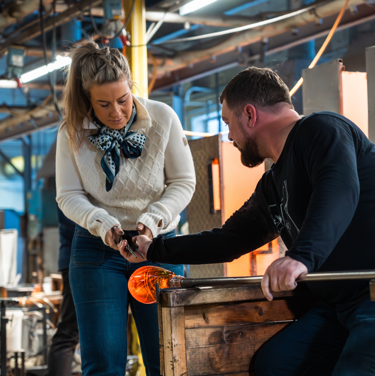 Woman and man working with molten glass in a workshop, shaping it with tools.