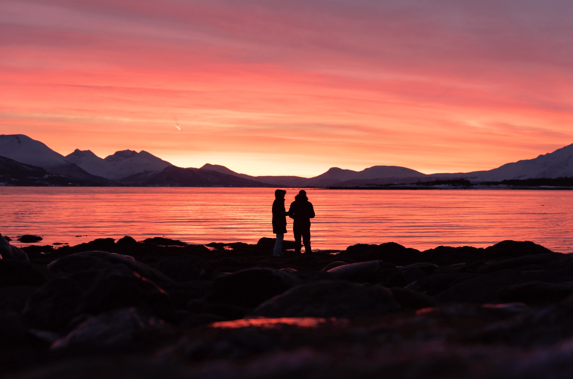 Silhouetted figures on a rocky shore watching a vibrant sunset over water and mountains.