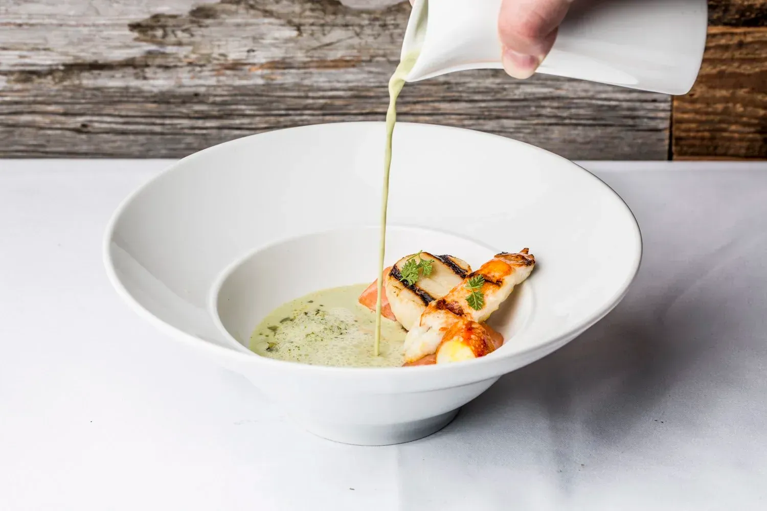 Soup being poured into a white bowl with seared scallops; wooden background.