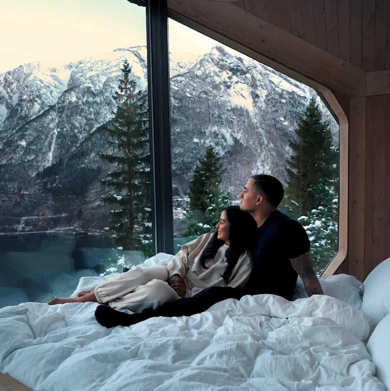 Couple on a bed looking at snowy mountains through a large window.