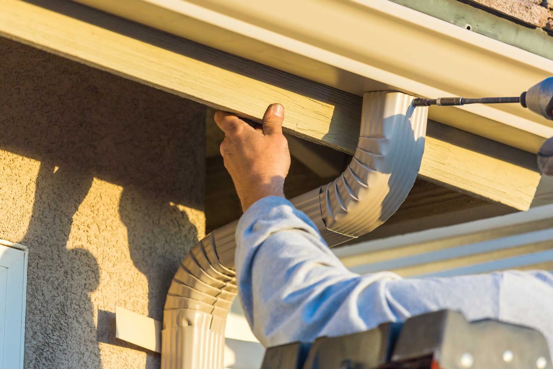 Worker installs an aluminum gutter to the wooden fascia of a house, outside of it and under the sun.