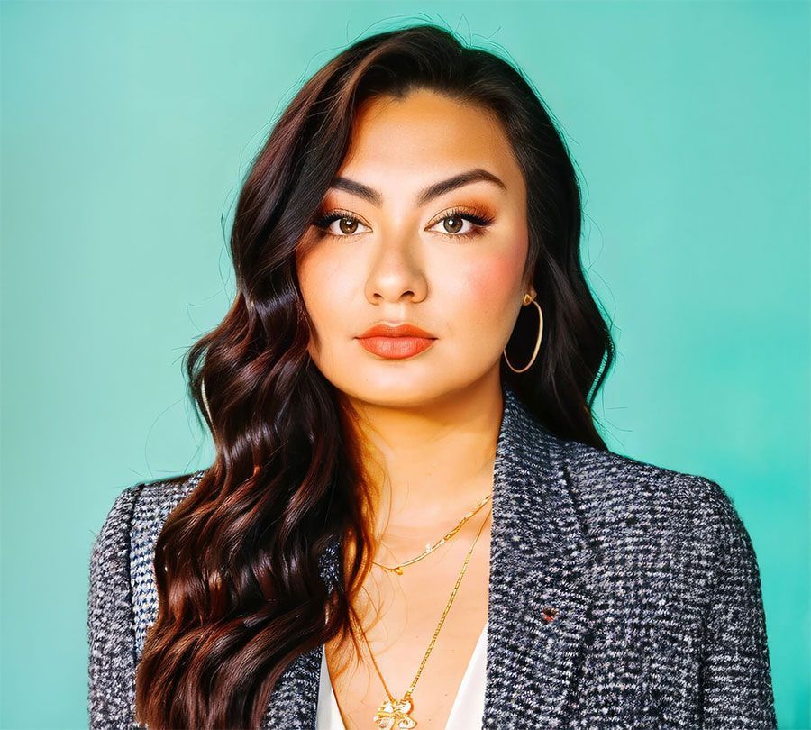A person with long, dark wavy hair wears a grey tweed blazer and gold layered necklaces against a turquoise background.