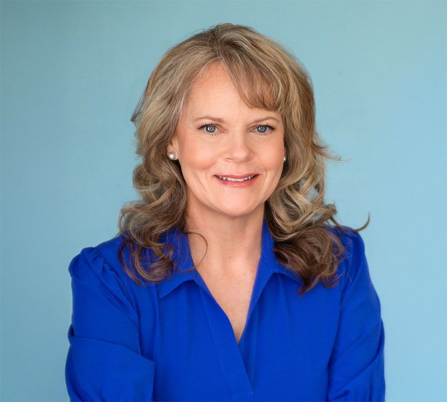A person with shoulder-length wavy hair wearing a bright blue collared shirt, smiling against a light blue background.