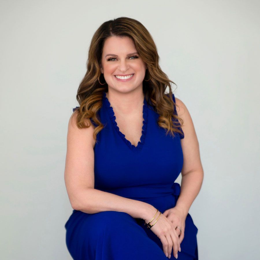 A smiling person with wavy brown hair wearing a sleeveless royal blue dress against a plain light background.