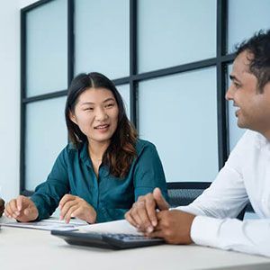 A woman and a man sitting at a desk in an office, facing each other and engaging in a professional discussion.
