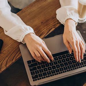 Hands typing on a laptop keyboard on a wooden table next to a glass of iced coffee.