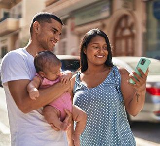 A person in a white shirt holds a baby while another person in a polka-dot dress looks at a smartphone on a city street.