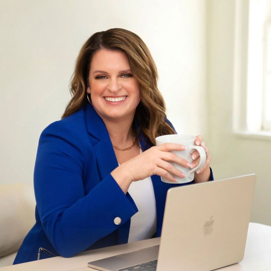 A person in a blue blazer smiles while holding a white mug and sitting in front of an open laptop in a bright office.