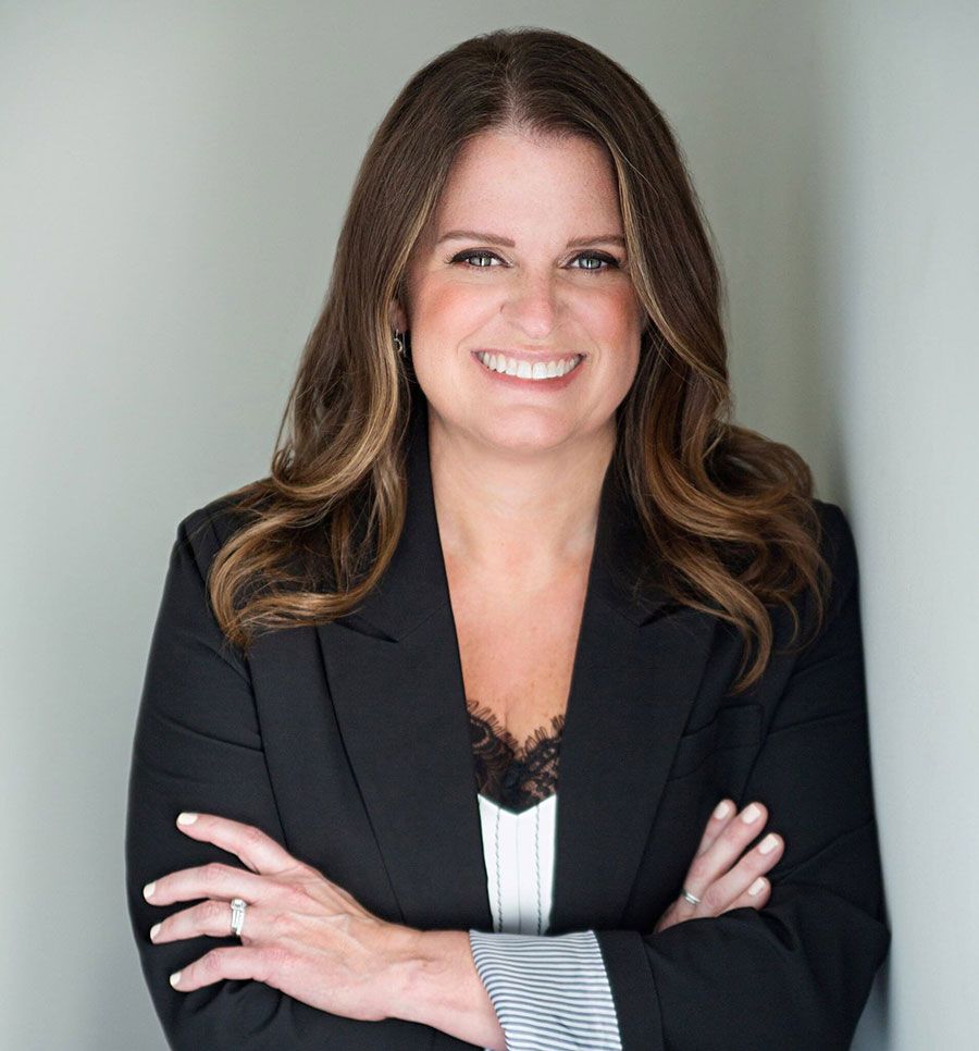A person with long brown hair, wearing a black blazer and white top, smiles with arms crossed against a neutral wall.