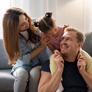 A family of three smiling together on a sofa, with a child reaching out to touch the adult's face.