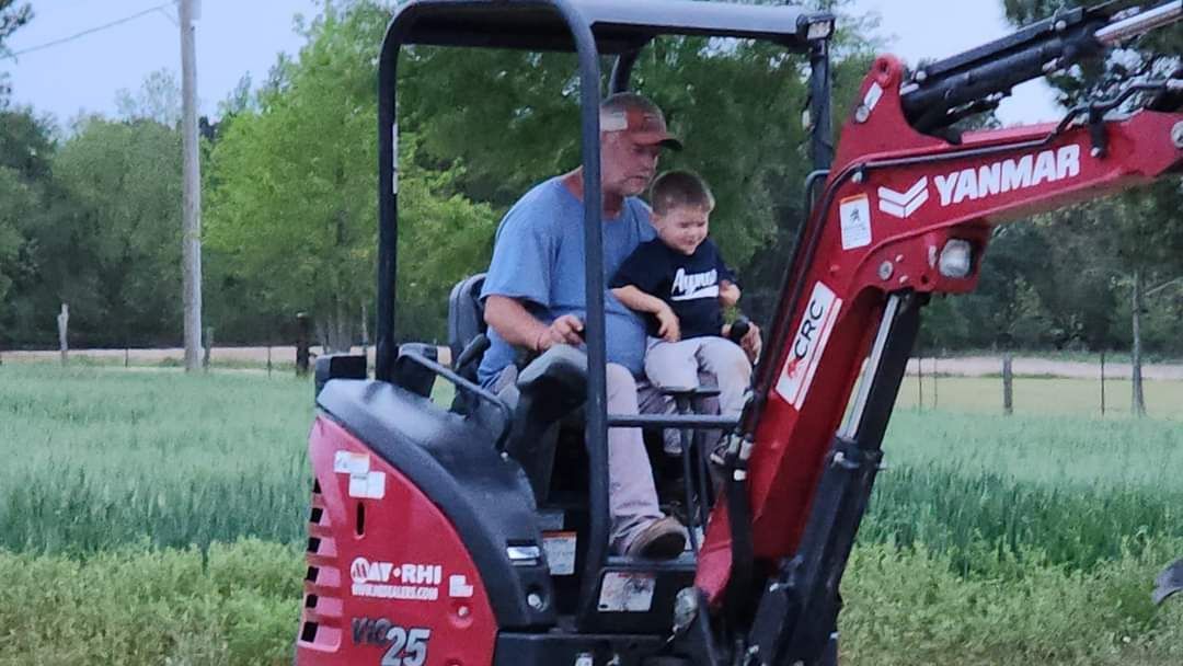 A man and a young child sit in the cab of a red Yanmar excavator in a grassy field.