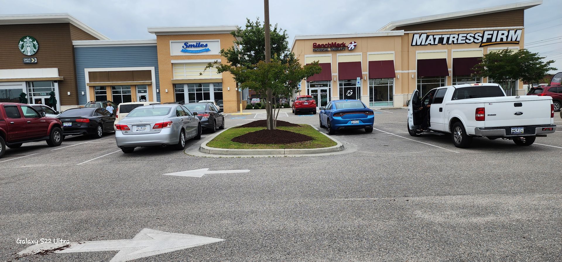 A parking lot with a Starbucks and other shops visible. Cars are parked, and a white truck has its door open.
