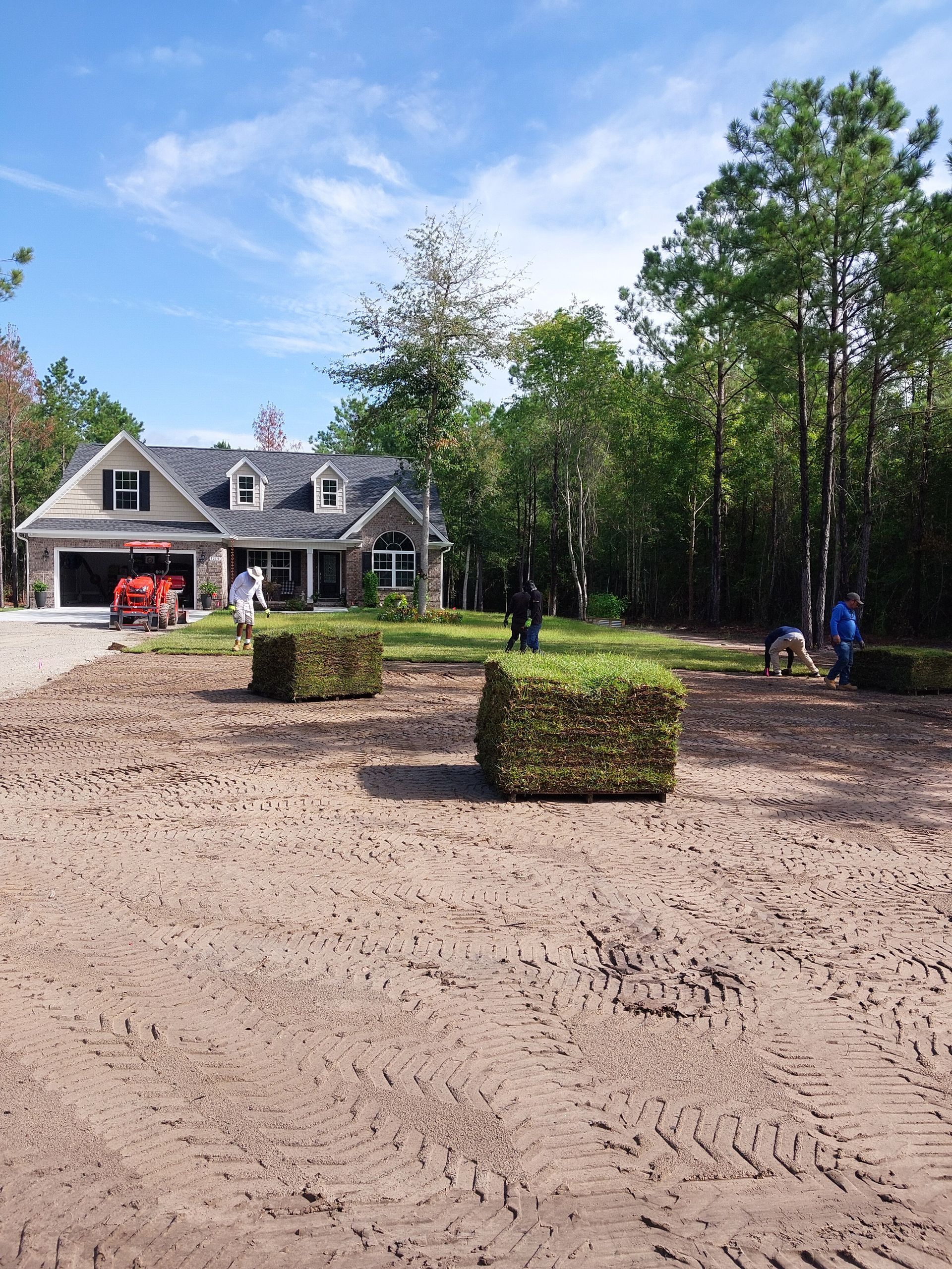 Workers installing sod in front of a house with a stone facade on a sunny day; several stacks of sod are visible.