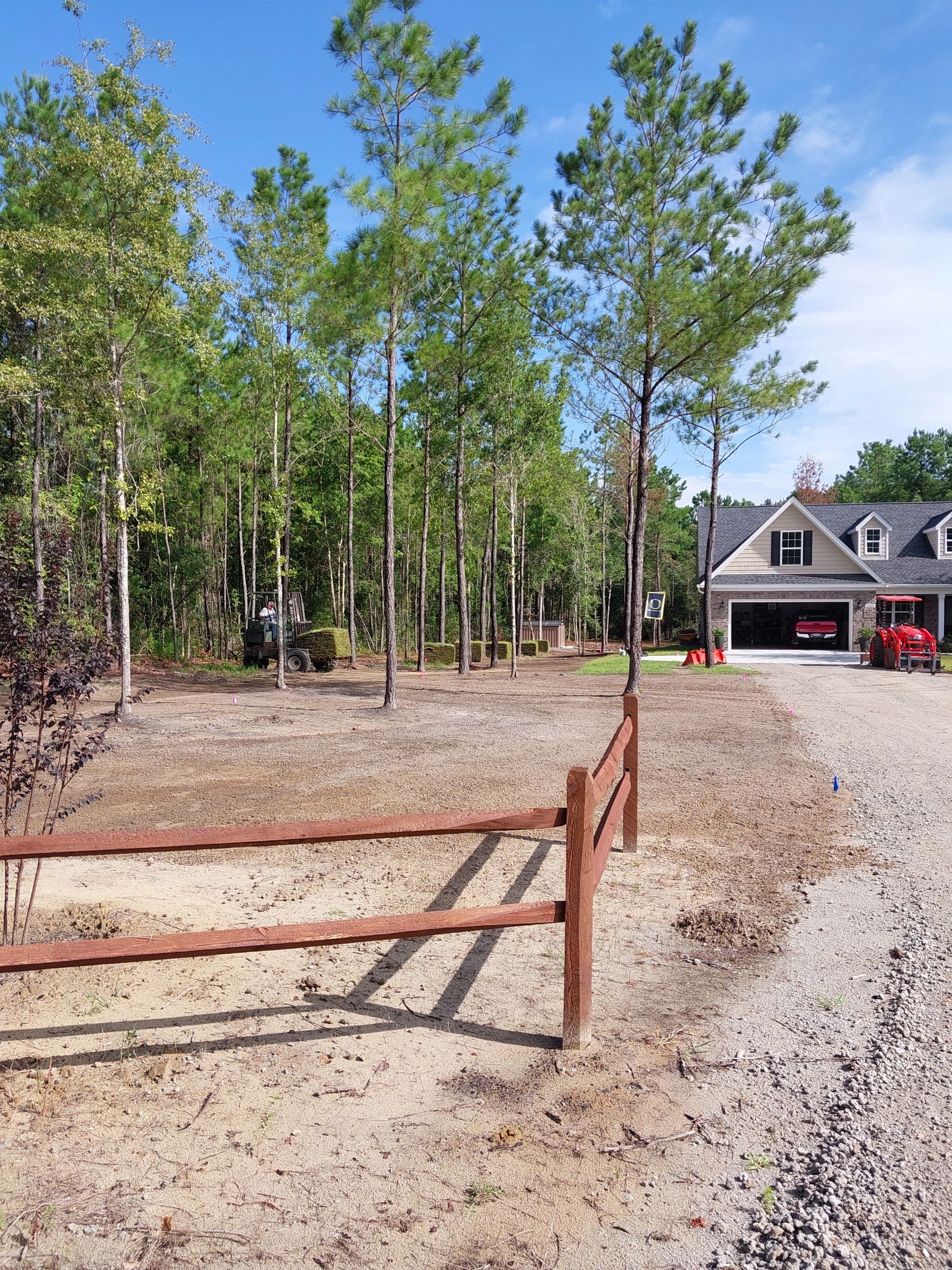 A gravel driveway leads to a house with a two-car garage. A wooden fence lines a cleared, dry area with young trees.