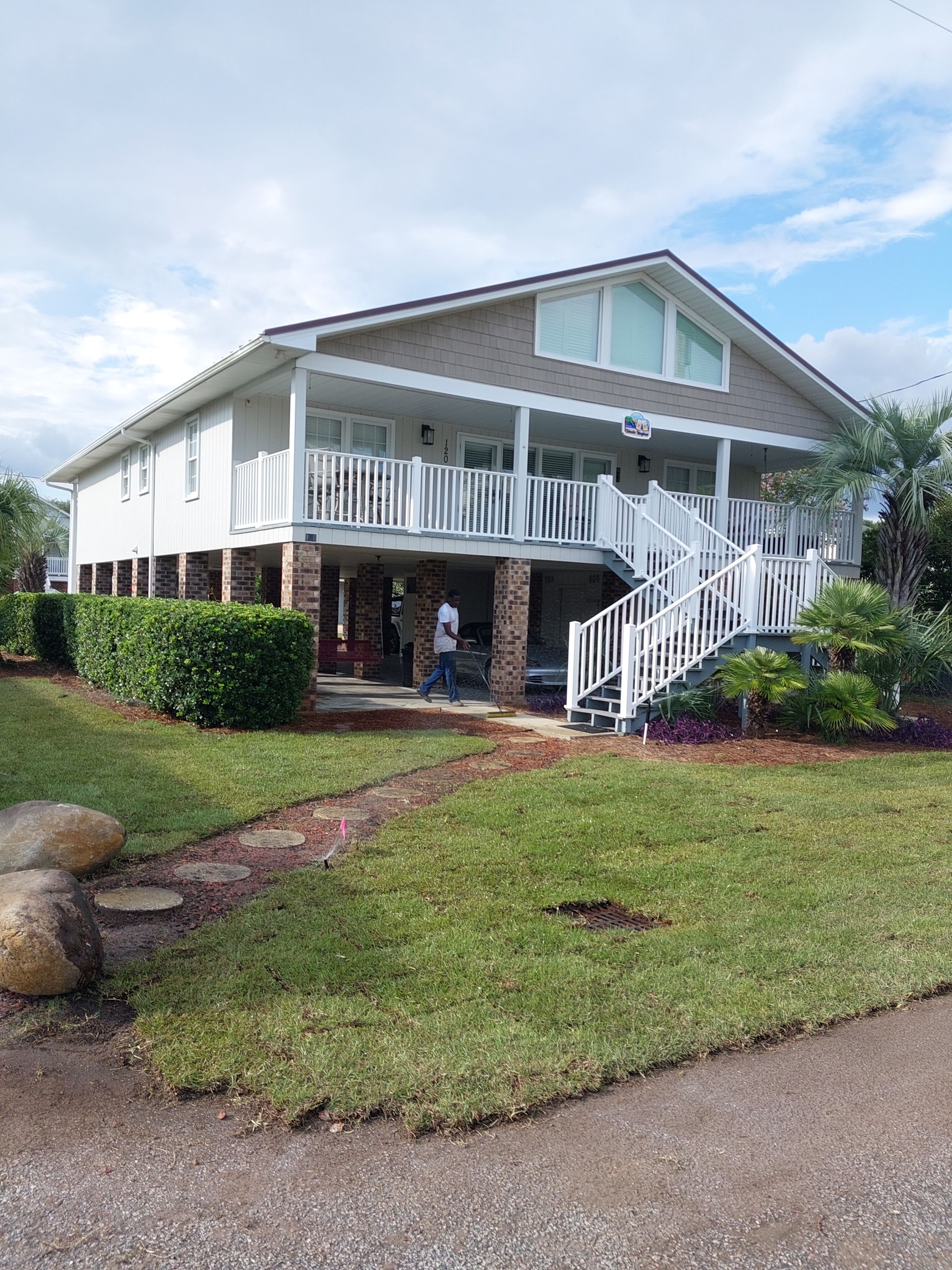 Two-story house on stilts with white railings and stairs, beige brick columns, and a small front yard with a person visible.