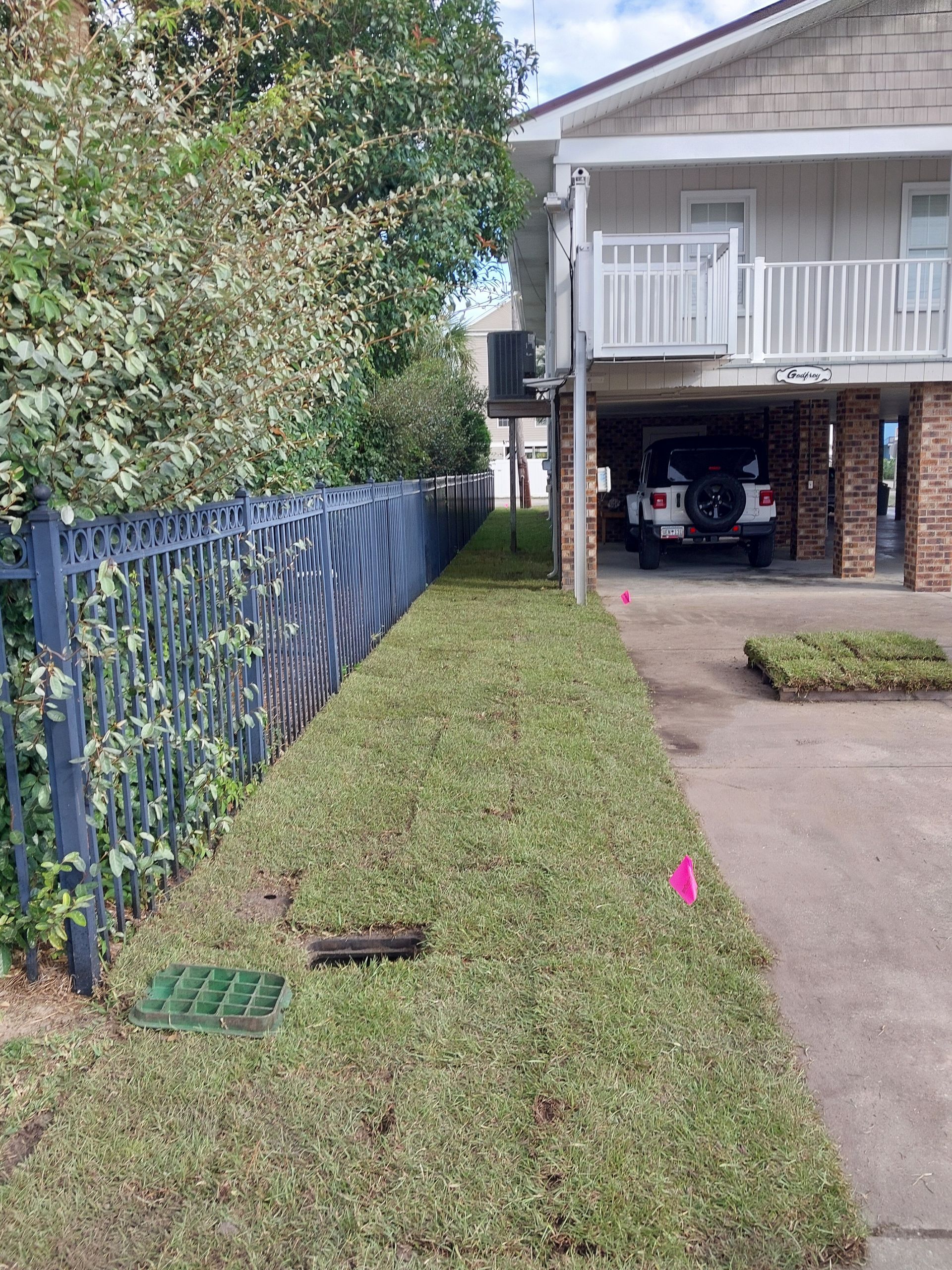 A narrow green area between a fence and a building, with cars parked beneath the building.
