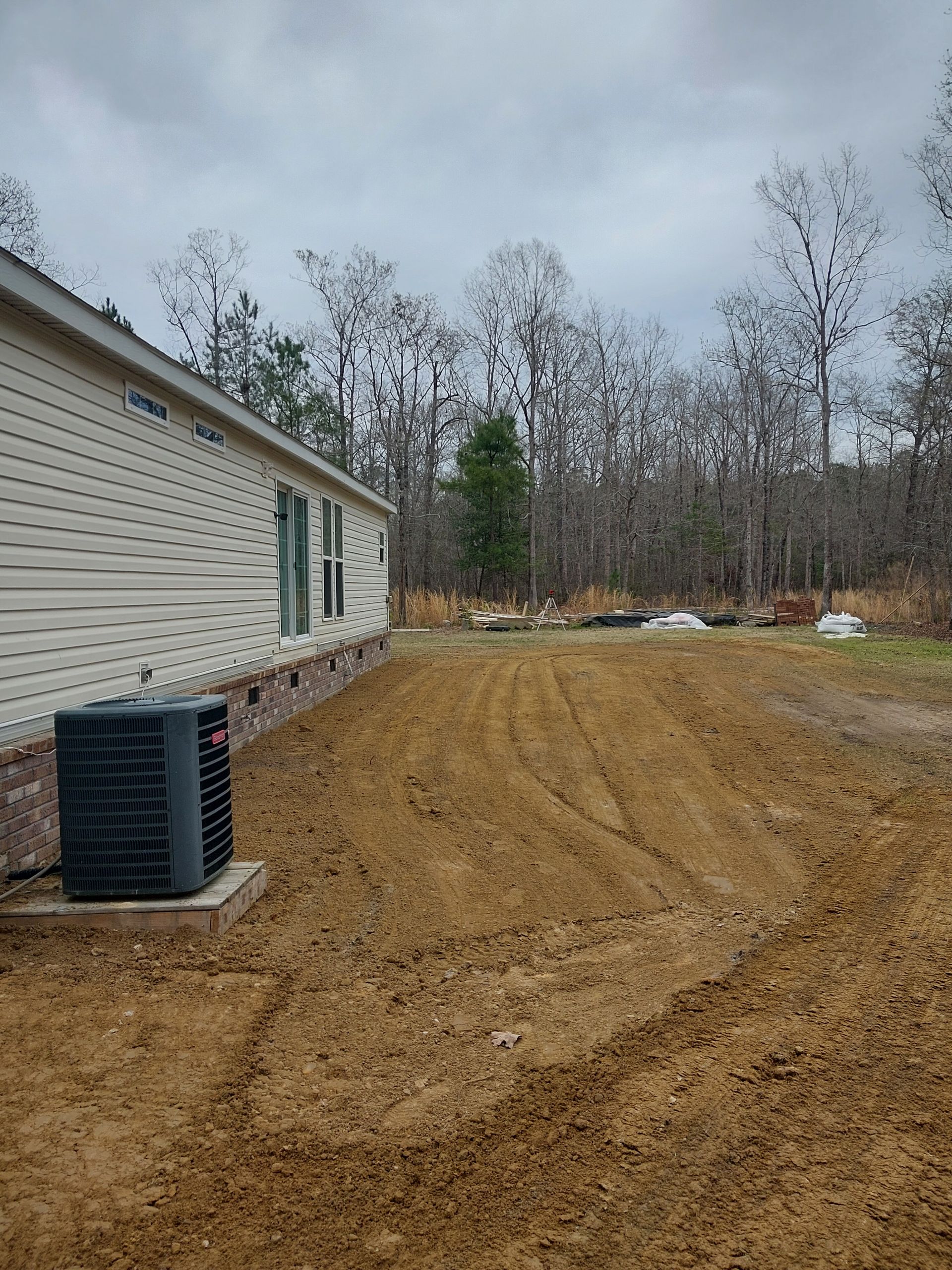 Side view of a beige house with an air conditioning unit in a muddy yard with tire tracks, surrounded by trees under a cloudy sky.