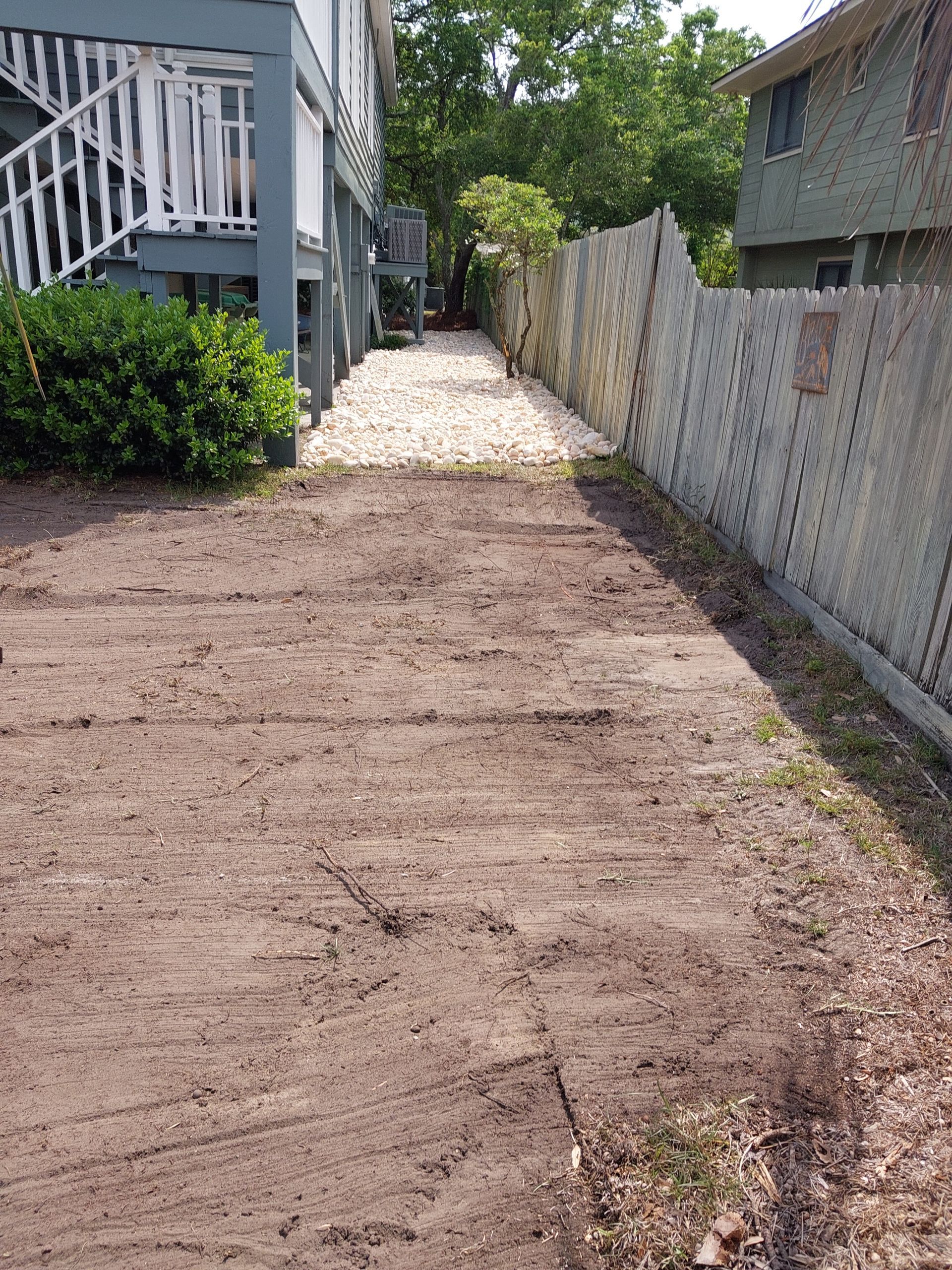 Dirt path leading between a house with a wooden deck and a weathered wooden fence. Pebbles cover a portion of the path.