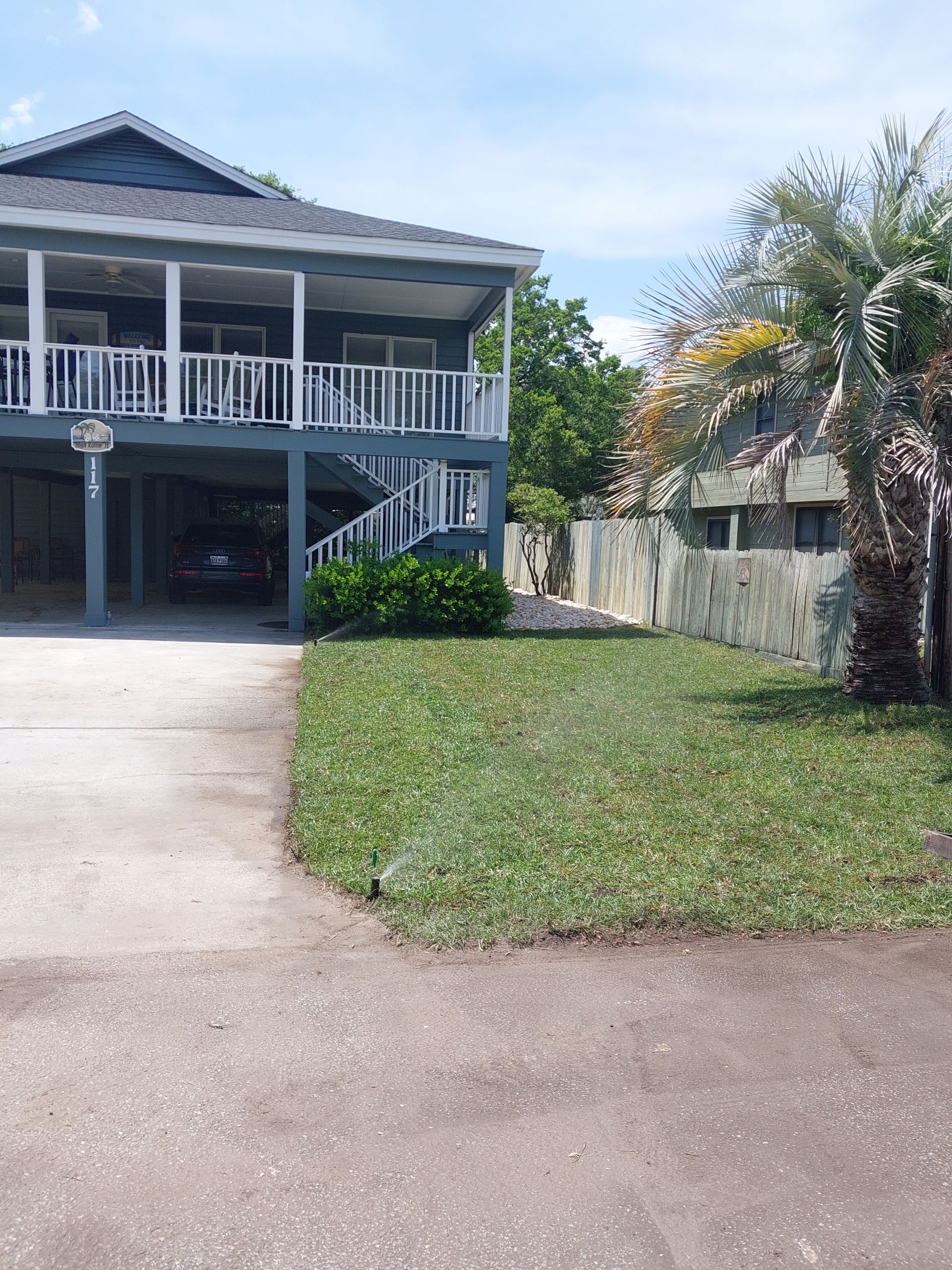 Two-story blue house with a porch and a driveway. A car is parked under the house. Green lawn and some palm trees.