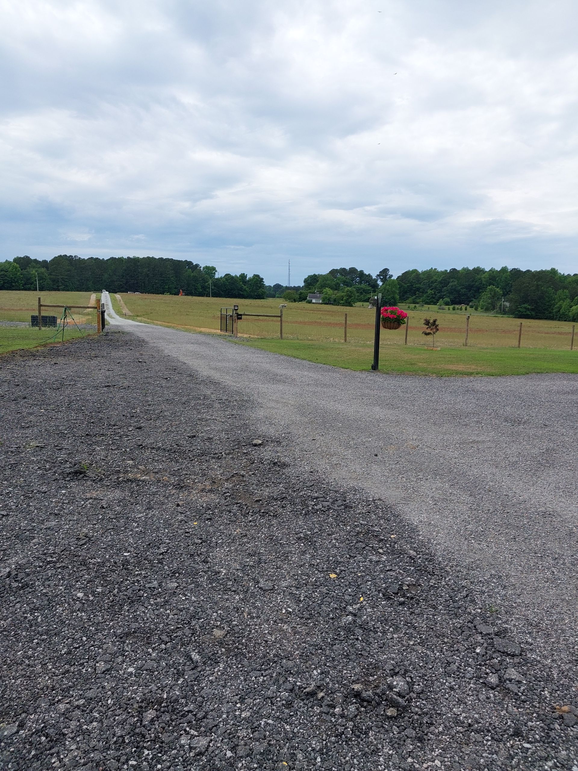 A gravel driveway leading to a field bordered by a wooden fence and treeline under a cloudy sky.