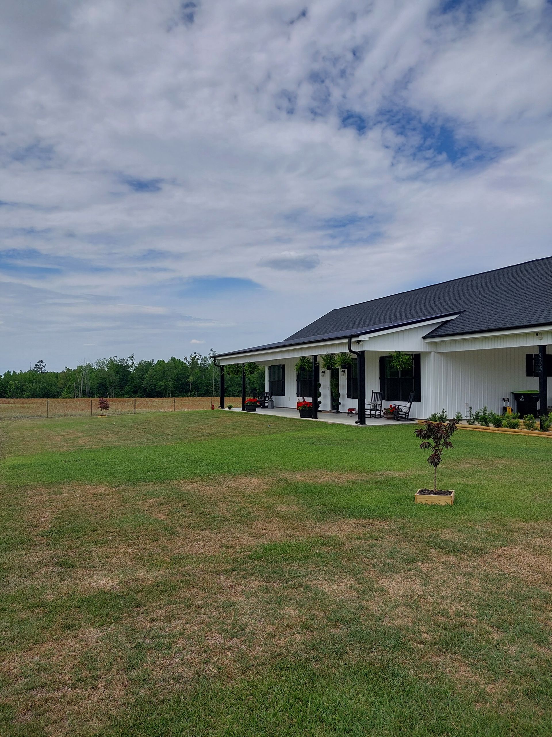 White house with a black roof and covered porch on a green lawn. A cloudy sky is above a treeline in the distance.