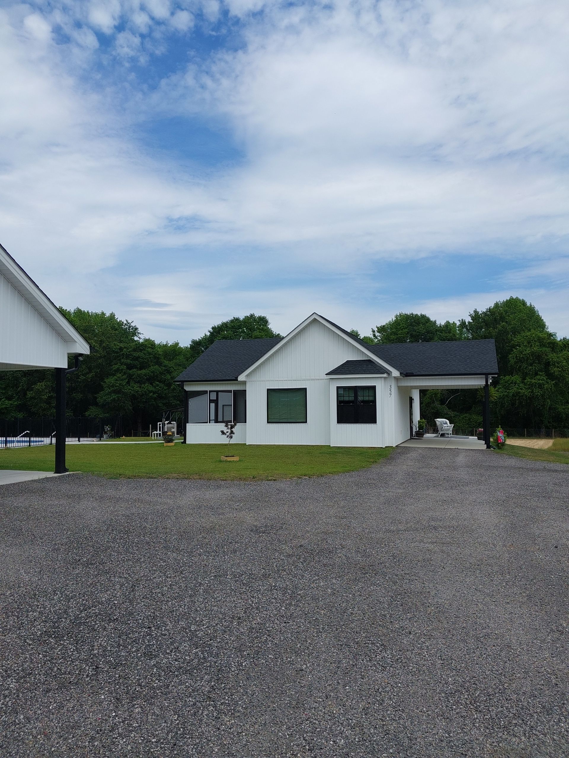 White house with black roof and windows, on a gravel driveway, with a clear blue sky and trees in the background.