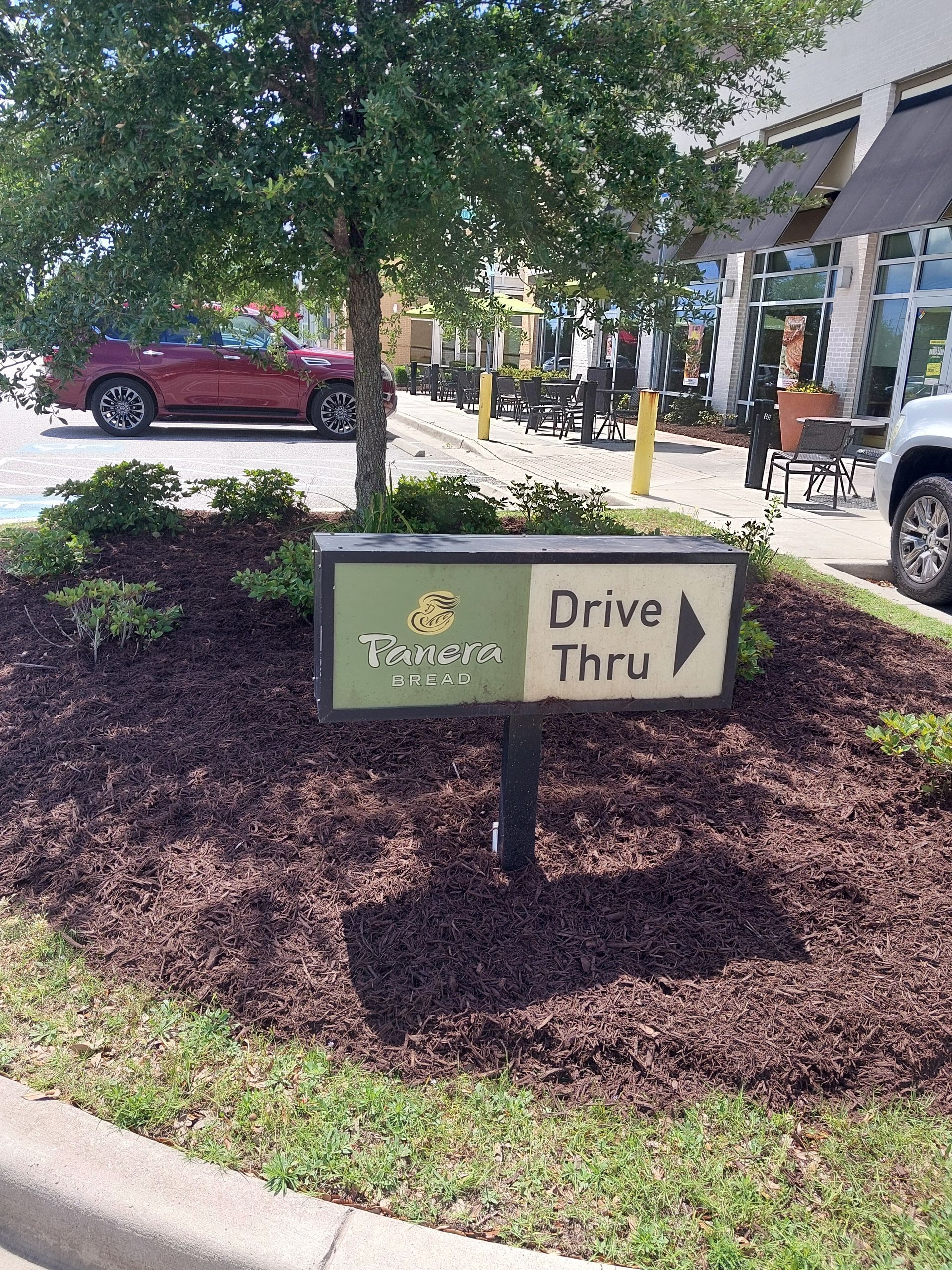 A Panera Bread drive-thru sign with arrow, surrounded by mulch and landscaping. A red car is parked in the background.
