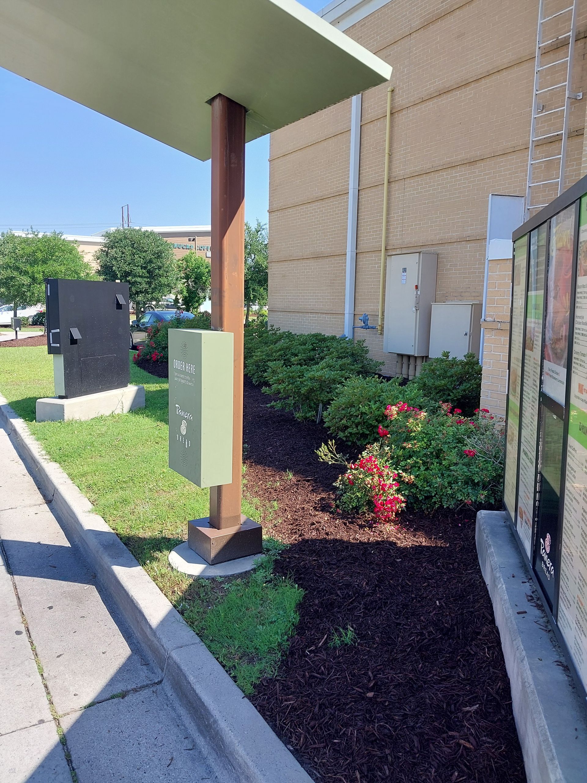 Green and brown electrical boxes near a drive-through canopy, beside a building with shrubbery and red flowers.