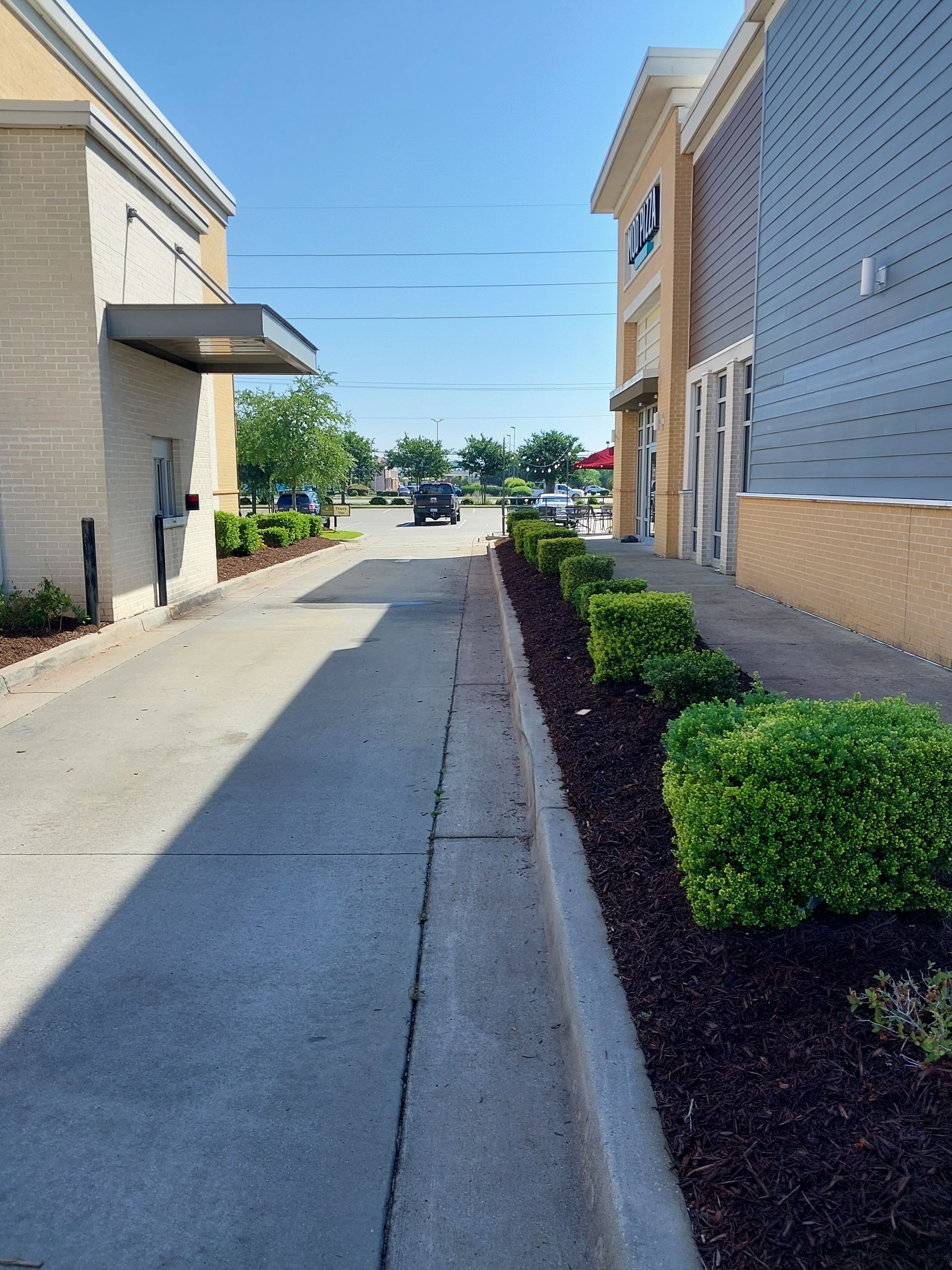 Drive-through lane with a building on each side. The lane is bordered by landscaping and leads toward other buildings and a bright sky.