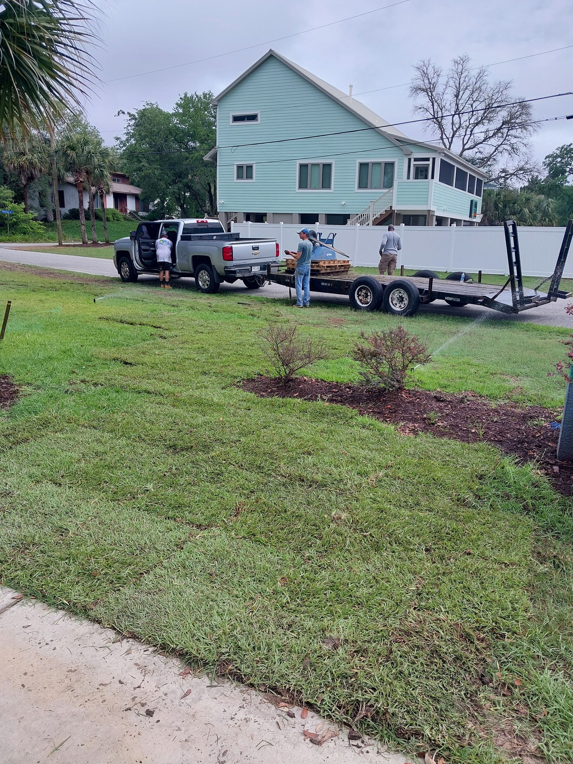 A silver pickup truck pulling a trailer with three people loading materials near a light blue house with a white fence.