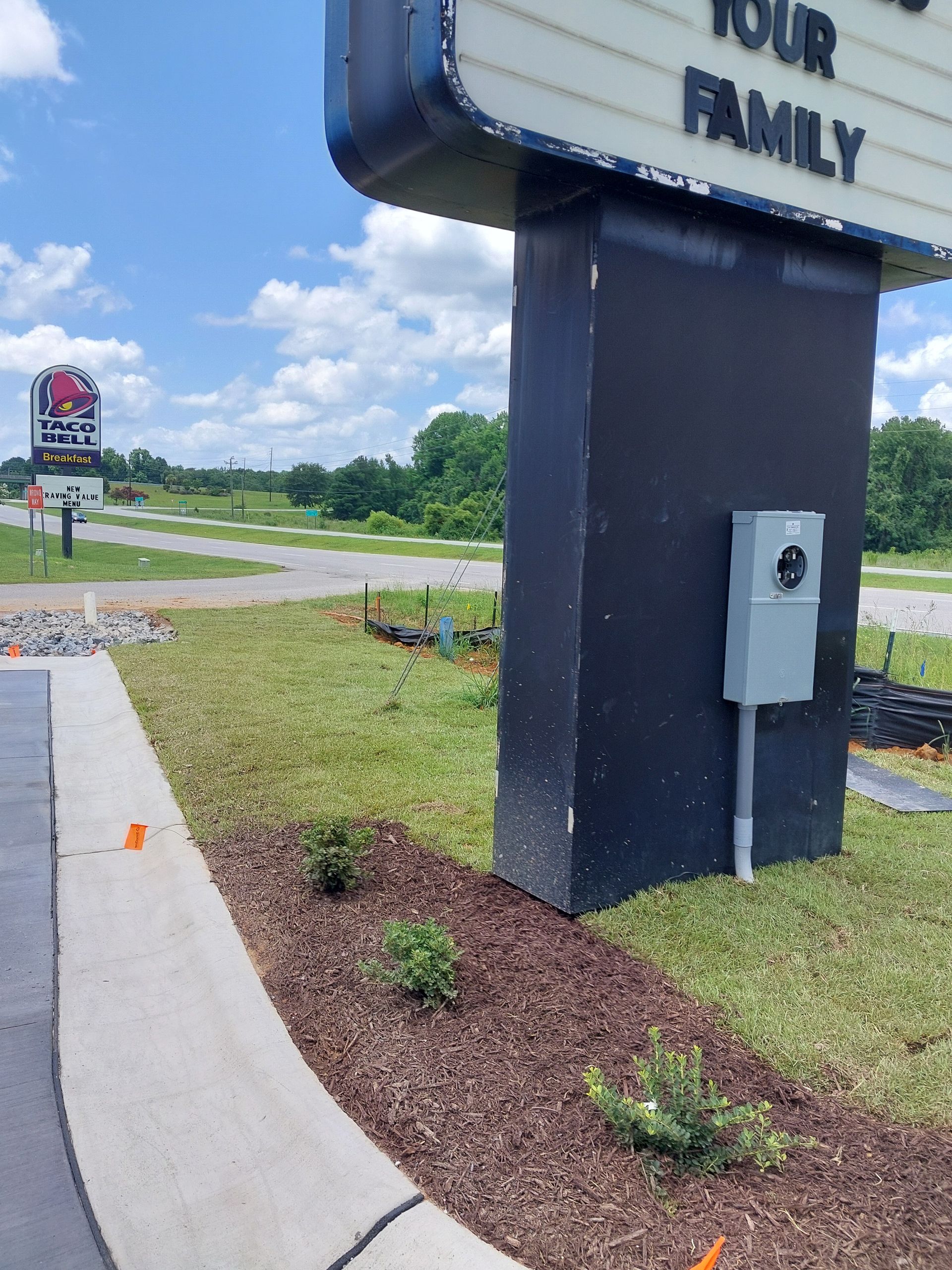 Signpost with electrical box near a curb, landscaped with bushes and mulch. In the distance, another sign is visible.