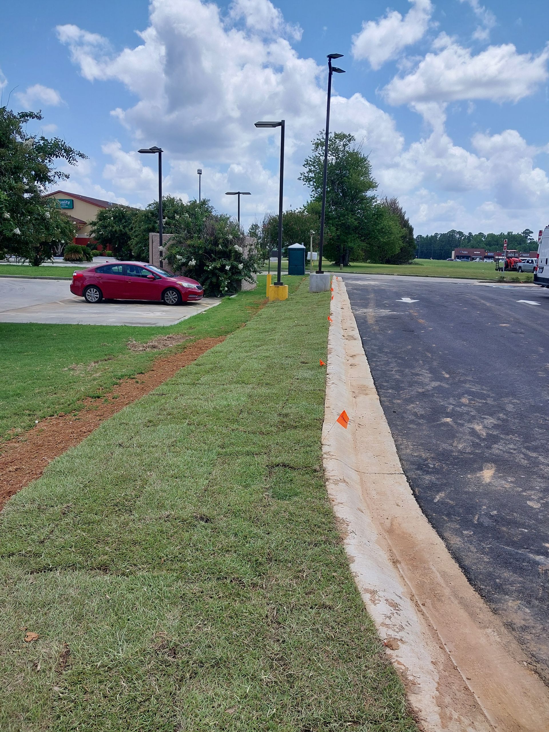 Green grass borders a parking lot with a concrete curb. A red car is parked near a building under a blue sky.