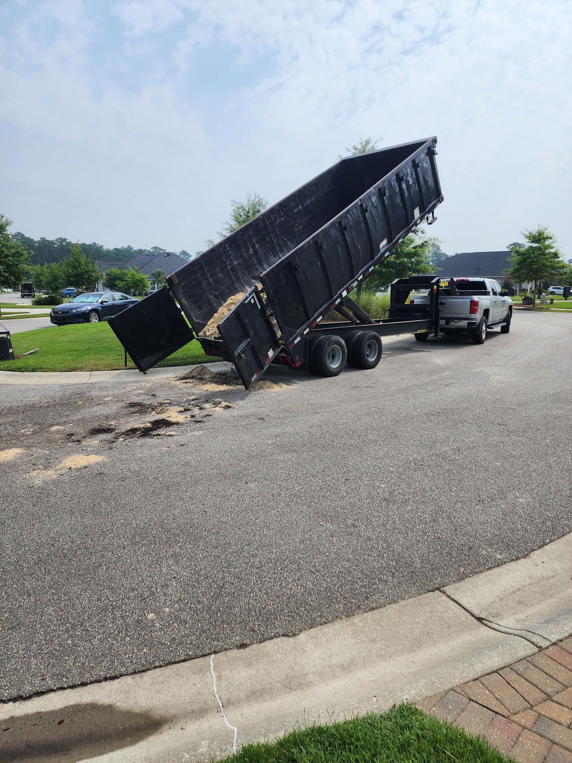 A pickup truck with a trailer dumping debris on an asphalt surface outdoors. The trailer is raised and dark, the truck is gray.